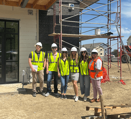 Six people wearing hard hats and safety vests stand together and smile at a construction site with scaffolding, building materials, and partially built houses in the background.