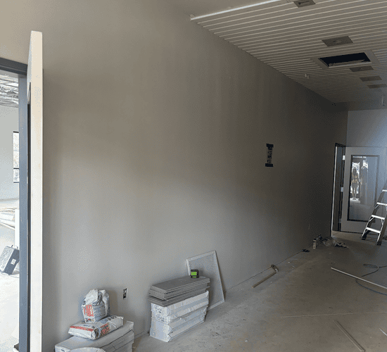 A construction site hallway with a newly painted beige wall, building materials and tools scattered on the floor, a ladder near glass doors, and ceiling panels partially installed.