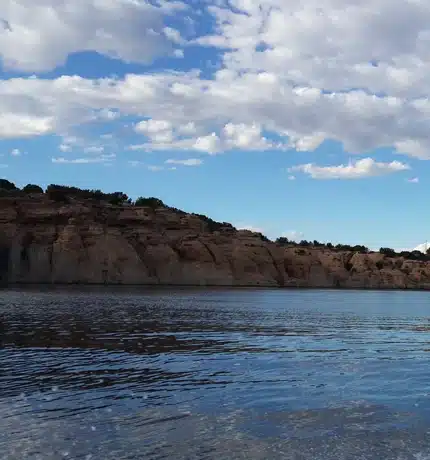 A calm body of water reflects the blue sky and clouds, with rocky, tree-topped cliffs lining the distant shore under scattered white clouds.