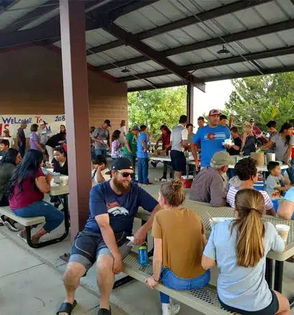 A large group of people socialize and eat together under a shaded outdoor pavilion. Some sit at picnic tables, while others stand and chat. A “Welcome” banner hangs on the wall in the background.