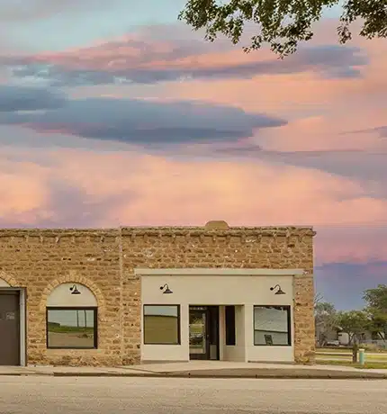 A small, tan brick building with large windows sits on an empty street under a colorful sunset sky with pink and purple clouds. A few trees are visible in the background.