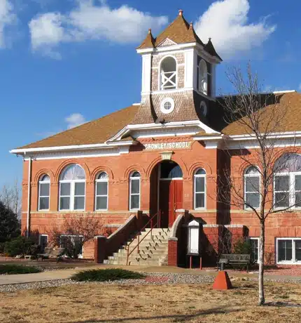 A historic red-brick building with arched windows, a central staircase, and a small white bell tower labeled CROWLEY SCHOOL, set against a blue sky with scattered clouds.
